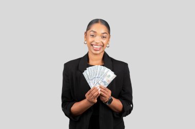 Bin yıllık Afro-Amerikan bir iş kadını şık bir siyah ceketle elinde neşeyle nakit para, finansal başarı ve kişisel kazancı somutlaştıran gri bir arka planda izole edilmiş bir yelpaze tutuyor.