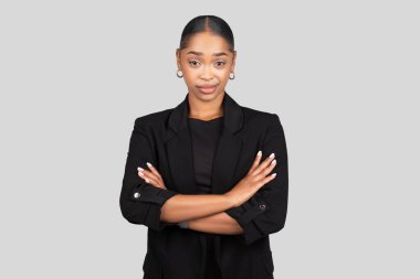 Milenyum kuşkucu Afro-Amerikan iş kadını kollarını kavuşturmuş şık, siyah ceketli, sakin, yaklaşılabilir bir gülümsemeyle gri bir stüdyo geçmişinde poz veriyor.