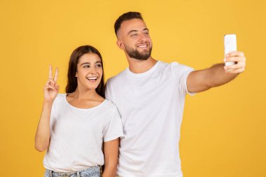 Neşeli beyaz bir kadın, gülümseyen bir adamla selfie çekerken barış işareti gösteriyor. Güneşli sarı bir arka planda, stüdyoda neşe ve dostluk anı yakalıyor.