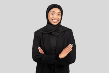 Kendine güvenen bin yıllık Afro-Amerikan iş kadını, tesettürlü, tesettürlü ve siyah ceketli, güven ve profesyonel duruş içinde kollarını kavuşturuyor.