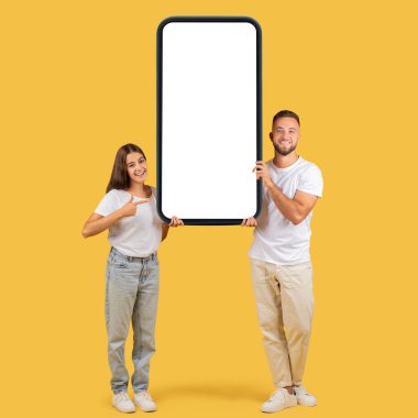 Gündelik giysiler içinde ışıl ışıl genç bir kadın ve erkek hardal sarısı arka planda reklam için ideal dev, boş bir akıllı telefon ekranına sahipler. Dikkat, iyi haber, web sitesi