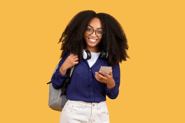 Sırt çantası taşıyan, gözlüklü siyahi bayan öğrenci cep telefonu kullanıyor ve gülümsüyor, sarı zemin üzerine kurulmuş, teknoloji ve öğrenci hayatının karışımını özetliyor.