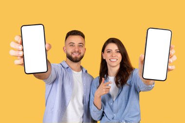 Gülümseyen adam ve kadın kameraya doğru boş ekranlı akıllı telefonları uzatıyor. Uygulama görüntüleme modelleri için mükemmel, sıcak sarı arka planda.
