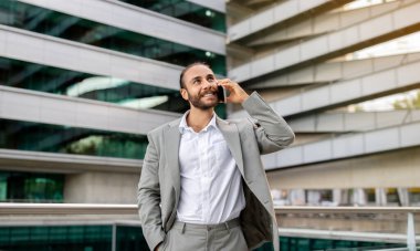 İş kıyafetleri içinde gülümseyen bir profesyonel cep telefonuyla konuşuyor ve yukarı bakıyor, mutlu genç iş adamı aktif kurumsal iletişimi somutlaştırıyor, dışarıda duruyor, uzayı kopyalıyor.