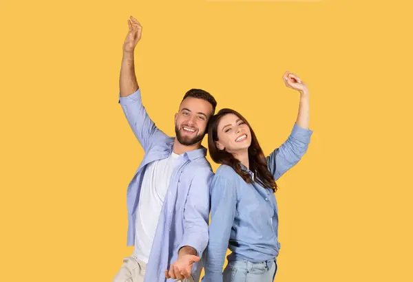 Man and woman joyfully dancing with arms raised high, smiling brightly, in casual blue attire, celebrating a moment on a cheerful yellow backdrop