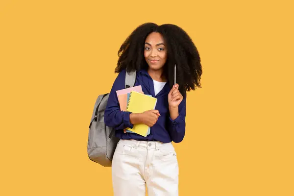 バックパックを運ぶカーリーヘアとノートブックとペンを保持しているスマートな若い黒人女性 晴れた黄色の背景に対して学校の準備ができて — ストック写真