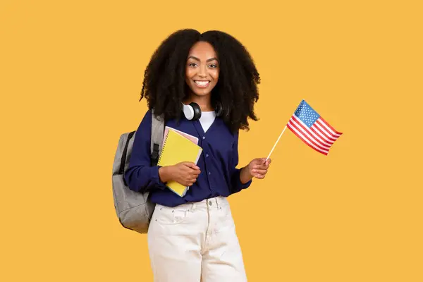 バックパックとコピーブックを装備した楽しい黒人女性の学生は 誇らしげに鮮やかな黄色の背景に対する米国の旗を保持し 愛国心と学術的な野心を象徴しています — ストック写真