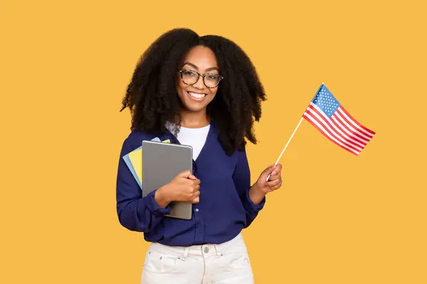 バックパックの黒人女性学生と黄色いスタジオの背景の上に米国の旗を示す本の束と笑顔 学校で英語を勉強するポジティブな女性 — ストック写真