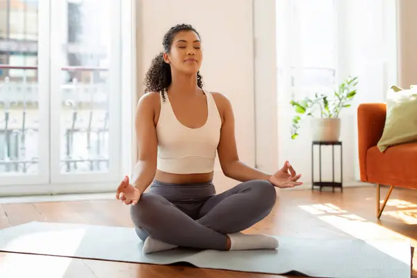 Peaceful fitness lady meditating on yoga mat, practicing mindfulness and meditation sitting in lotus position in modern living room interior in the morning, closing eyes while breathing