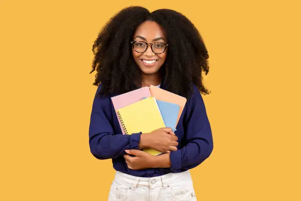 カーリーヘアとメガネのインテリジェントな若い黒人女性 喜んでカラフルなノートブックのスタックを抱擁し 黄色の背景に対するクラスの準備 — ストック写真
