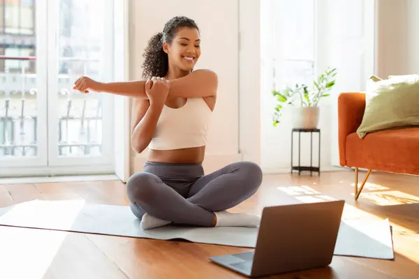 Happy fitness lady looking at laptop during online pilates workout, stretching arm near computer, sitting in lotus position on yoga mat in modern living room in the morning