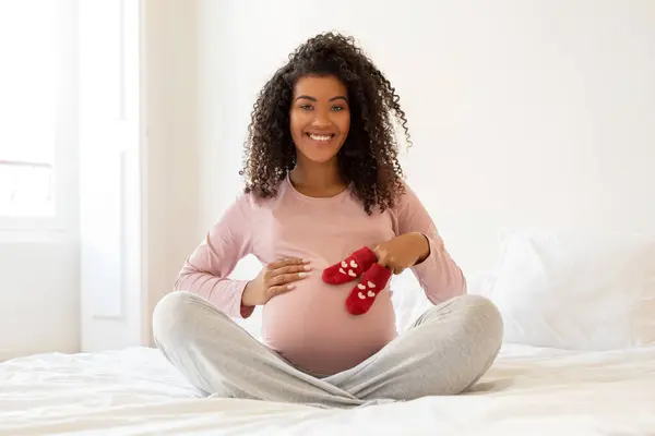 マタニティ コンセプト 黒い妊娠中の女性は腹を抱き 自宅でベッドに座っている間 小さな赤ちゃんの靴下を手に持っています 幸せなアフリカ系アメリカ人の期待する女性は妊娠時間を楽しんで スペースをコピーします — ストック写真