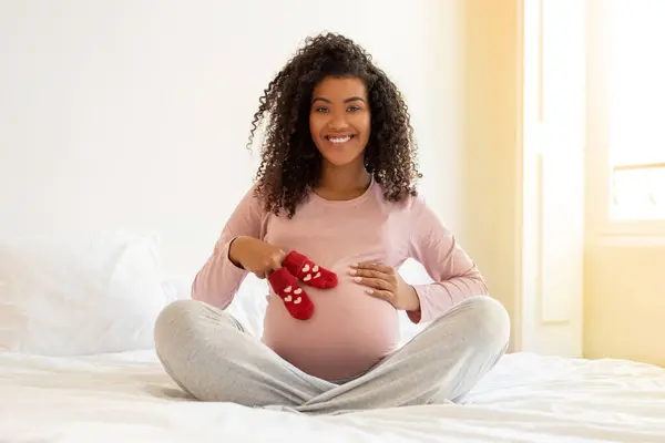 母性のために準備する黒い妊婦 腹の近くに小さな赤ん坊のソックスを保持し ベッドに座って笑顔でアフリカ系アメリカ人女性 マタニティを楽しむ コピースペース — ストック写真