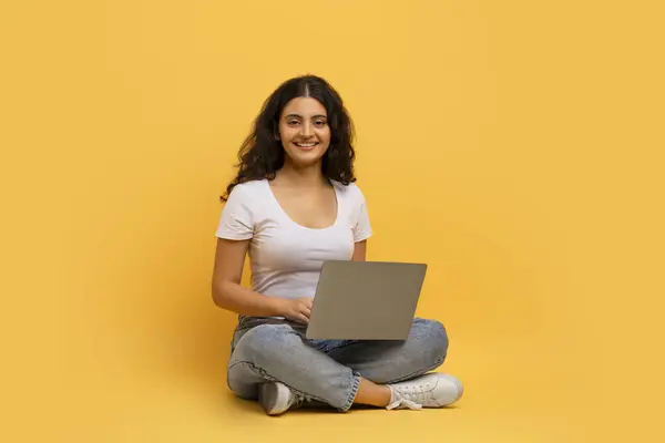 ハッピーな若いヒンドゥーの女性フリーランサーは コンピュータのラップトップのキーボードに入力し オンラインで作業し 黄色のスタジオの背景に隔離された床に座っています リモートジョブ コピースペース — ストック写真