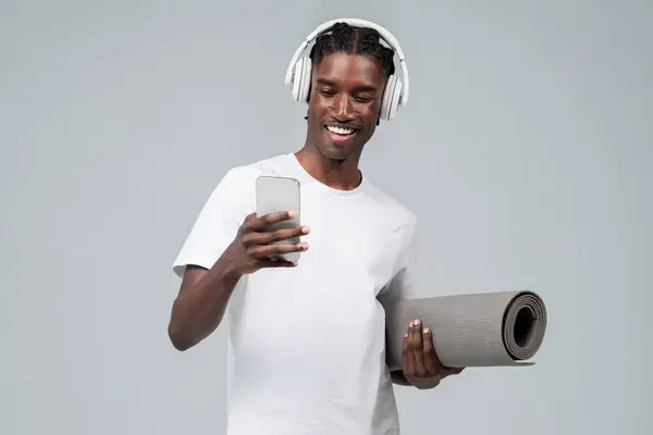 スマートフォンやヨガマットを保持するワイヤレスヘッドフォンを身に着けている楽しい黒人男性 音楽やフィットネスアプリに従事する陽気なハンサムなアフリカ系アメリカ人男性 灰色のスタジオの背景に立って — ストック写真