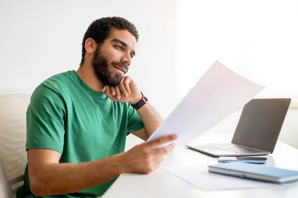 stock image Contented middle eastern young man with a beard, wearing a green t-shirt, looks at a document while sitting at a bright work desk, with a laptop and smartphone nearby at home