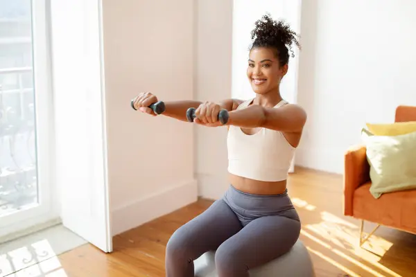 Fit woman sitting on balance ball and lifting weights exercising with dumbbells in her modern living room at home, having workout in the morning. Strength and bodybuilding training