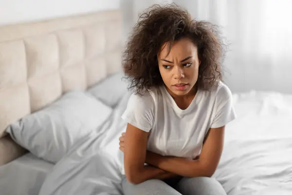 Depressed African American Young Lady Hugging Herself Suffering From Depression And Feeling Bad, Having Stomachache Problem Or Painful Periods, Sitting In Modern Bedroom At Home