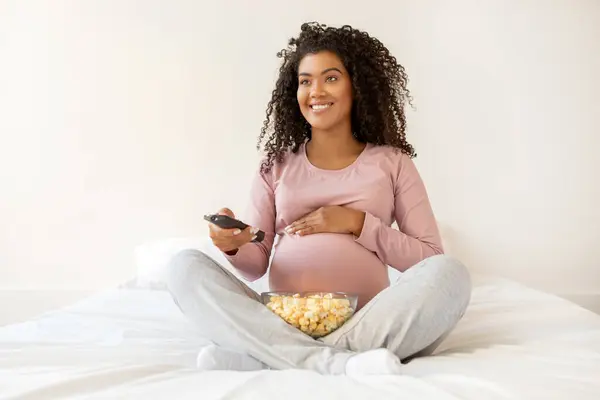 Happy Black Pregnant Lady Relaxing At Home, Watching Tv And Eating Popcorn In Bed, Smiling Expectant African American Woman Holding Remote Controller, Resting In Cozy Bedroom, Copy Space