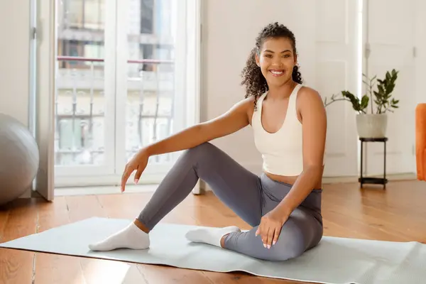 Fit young woman in sportswear sitting on mat smiling to camera, posing ready for morning yoga or fitness workout at home interior. Healthy active lifestyle and sport routine