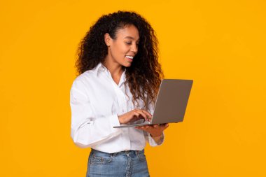 Mutlu Afro-Amerikan bayan dizüstü bilgisayarını kullanarak online iş yapıyor, internet reklamları yapıyor, sarı stüdyo arkaplanı üzerinden not defterine göz atıyor. Serbest meslek.