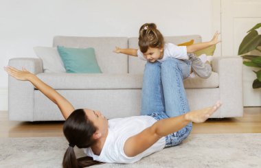 Anne ve küçük kızı evde birlikte eğleniyor, annesi kollarını açmış sırt üstü yatıyor, keyifli çocuğunu bir uçak oyununda kaldırıyor, oturma odasında oynuyor ve gülüyor.