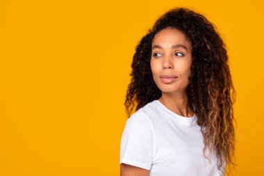 Afro-Amerikalı genç bir kadının portresi, düşünceli bir şekilde, parlak sarı stüdyo arkaplanına karşı poz vererek, beyaz t-shirt giyen, mesaj reklamı için boş alana bakıyor.