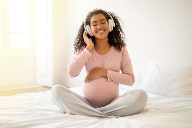 Mutlu siyah hamile kadın yatakta oturuyor, müzik dinliyor ve karnına dokunuyor, gülümsüyor kablosuz kulaklıklı Afro-Amerikan kadın yatak odasında dinleniyor, hamilelik zamanının tadını çıkarıyor,