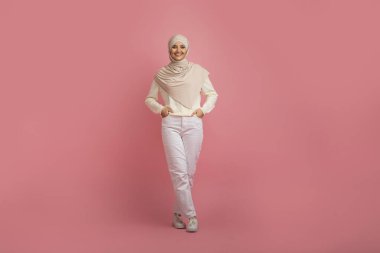 Neşeli Arap Müslüman kadın zarif bir şekilde canlı pembe stüdyo arka planında yürüyor, mutlu kadın pozitiflik ve güven yayıyor, geleneksel kültürü çağdaş tarzla harmanlıyor.