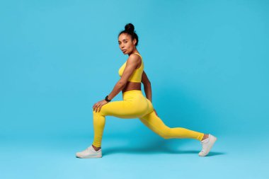 Egzersiz eğitimi. Afro-Amerikalı fitness bayan öne doğru hareket eden bacakları esnetiyor, mavi stüdyo arka planında sarı bir elbise giyiyor, dayanıklılık ve sağlığın vücut bulmuş hali..