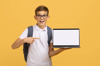 Gözlüklü hevesli öğrenci boş beyaz dizüstü bilgisayarı işaret ediyor. Sırt çantalı mutlu genç erkek çocuk sarı arka planda duruyor. Dijital öğrenmeyi ya da internet eğitimini sembolize ediyor.