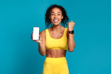 Antrenman uygulamaları ve aletler. Beyaz ekranlı akıllı telefon, bileğinde spor ayakkabı, sarı spor kıyafetlerle mavi stüdyo arka planında poz veren mutlu siyahi kadın. Model