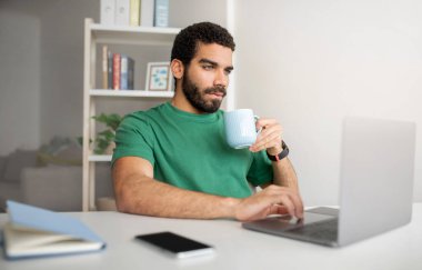 Yeşil tişörtlü bir adam beyaz bir masada dizüstü bilgisayarda çalışıyor, mavi bardaktan kahve içiyor, arka planda defter, akıllı telefon ve kitaplık olan bir ofis ortamında.