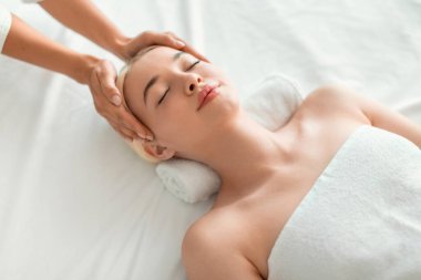 Yüz geliştirme ve gençleştirme. Gözleri kapalı olan Avrupalı sarışın kadın, kaplıca merkezinde cilt bakımı görüyor ve yüz masajı yaptırıyor, beyaz havluya sarılmış olarak yatıyor.
