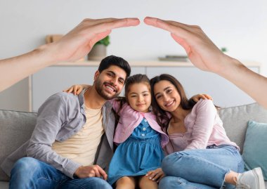 Mutlu ırklar arası aile babası, anne ve küçük kızı evdeki kanepede kucaklaşarak poz veriyorlar. Orta doğulu kız ve ebeveynler el altında oturup onlara çatı yapıyorlar. Sigorta konsepti