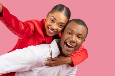 Coşkulu genç siyah çift selfie çekiyor, kırmızı bluzlu kadın erkeklerin arkasına yaslanıyor, adam pembe arka planda kadını sırtında taşıyor.