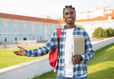 Elinde dizüstü bilgisayarı ve kitapları olan, üniversite kampüs parkında gelişigüzel dikilen, uluslararası üniversite öğrenim programına davet ve reklam veren Afro-Amerikalı genç bir öğrenci.