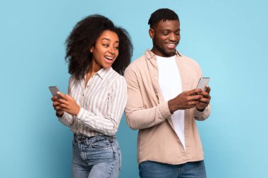 Neşeli genç siyah çift, arka arkaya, akıllı telefonlarıyla derinden ilgileniyorlar, rahatlatıcı açık mavi arka plana karşı birlikte neşeli anı paylaşıyorlar, modern iletişimi somutlaştırıyorlar.