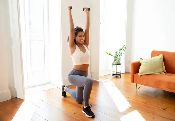 stock image Cheerful athletic woman standing in lunge position and raising arms holding dumbbells, having bodybuilding workout in modern living room interior. Home fitness concept