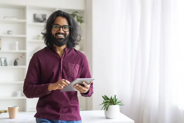 手元にデジタルタブレットを持つハンサムなインドの男性フリーランスの肖像自宅のオフィスでデスクの近くに立って 千年紀の男性ホールディングタブコンピュータとカメラで微笑む 現代の技術を楽しんで — ストック写真