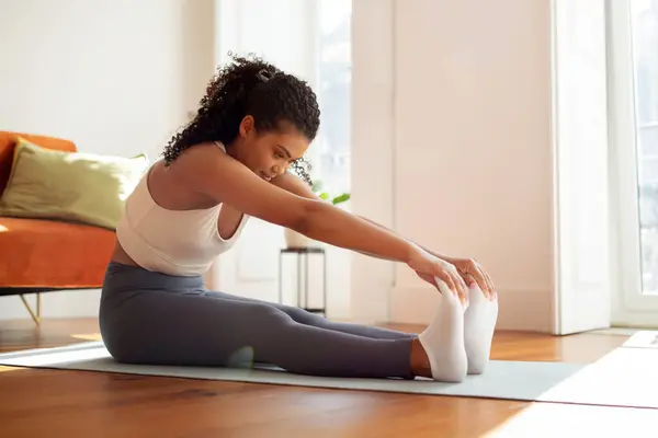 Sporty lady seated on yoga mat practicing pilates performing toe touch aerobics stretching exercise, showcasing her physical strength and flexibility in living room indoors.