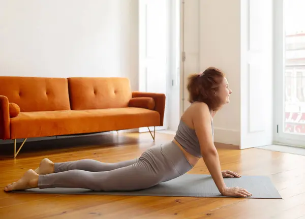 Senior lady training at home, making Cobra pose on yoga mat, sporty elderly woman exercising in living room interior, older female enjoying active retirement and healthy lifestyle, copy space