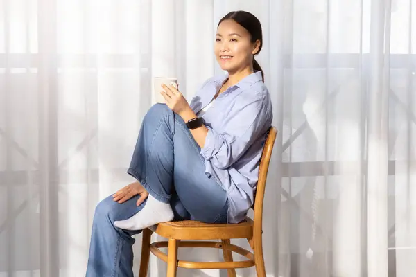 Happy attractive millennial chinese woman in casual clothing sitting on chair next to window at home, drinking tea or coffee, looking at copy space and smiling, sunshine. Domestic relaxation