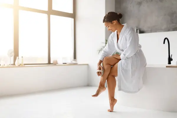 身穿白色浴衣的黑人妇女用干枯的身体刷擦去腿毛 快乐的非洲裔美国女性坐在现代通风浴室窗户边的浴缸上 复制空间 — 图库照片