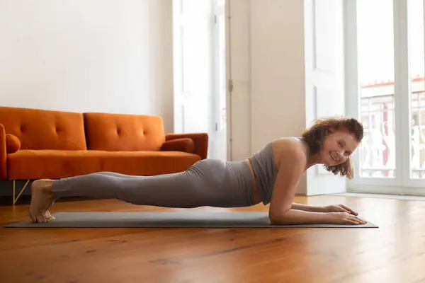 Senior woman with bright smile maintaining plank pose while training at home, beautiful elderly lady demonstrating strength and flexibility in her domestic workout routine, smiling at camera