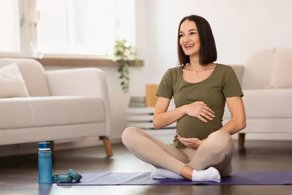 Healthy Pregnancy Workout. Pregnant woman expecting baby sits in lotus pose on floor at home, embracing her belly while engaging in prenatal yoga session, focusing on wellbeing