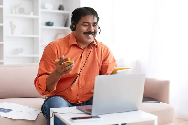 Online education, e-learning. Mature indian man studying online from home, sitting on couch, using headset and laptop, taking notes while have video call with teacher tutor, copy space