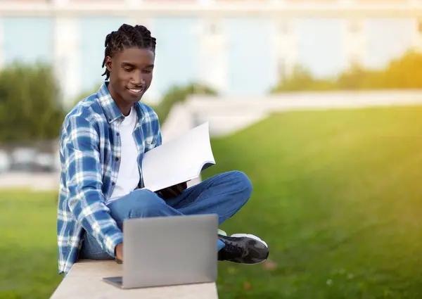 African American student guy websurfing on laptop holding workbooks and websurfing, studying sitting on grass outside university campus, immersed in digital learning leisure. Copy space
