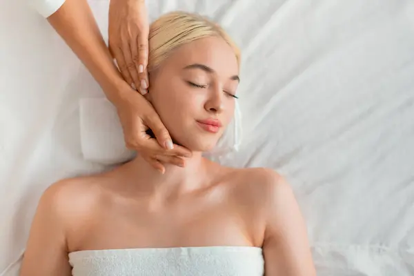 Top View Of Young Blonde European Lady Enjoying Facebuilding Massage Indoors. Woman Receiving Facial Beauty Procedure, Portrait Shot From Above. Pampering And Relaxation