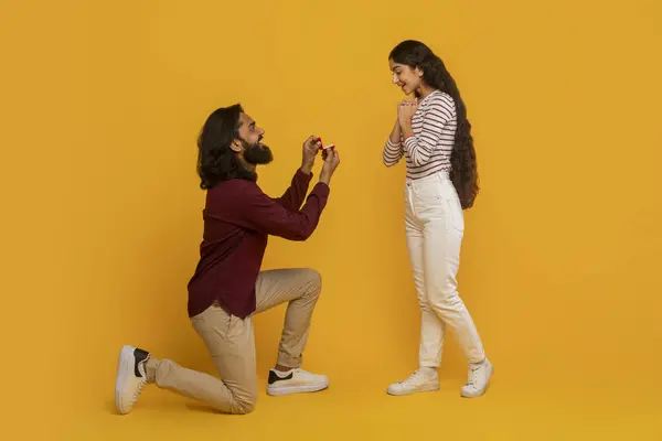 美しいハンサムなインドの男性は 彼のかなり興奮したガールフレンドに結婚の提案を行い 1つの膝の上に立って リング 黄色の背景を持つボックスを保持します エンゲージメント — ストック写真